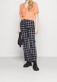 Orange stickad kortärmad cardigan, svart- och vitrutiga byxor, svarta klumpiga ankelskot, håller en liten svart väska.