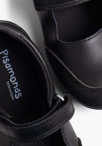 Pantofi Mary Jane din piele neagră cu curea elastică, panou texturat și finisaj neted. Logo-ul spune "Pisamonas, Hecho en España."
