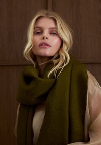 Écharpe en laine vert olive drapée autour du cou, présentant une texture douce et un design ample et surdimensionné. Fond neutre avec des panneaux en bois.