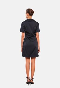 Schwarzes kurzärmeliges Kleid mit Kragen, aus einem glatten Stoff gefertigt, in einer figurbetonten Silhouette gestaltet, endet auf halber Oberschenkelhöhe, kombiniert mit schwarzen High Heels.