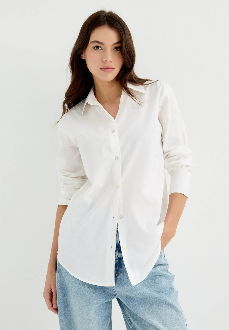Camisa blanca de botones con mangas largas, dobladillo redondeado y textura de tela. Combinada con vaqueros de mezclilla sueltos de color azul claro.