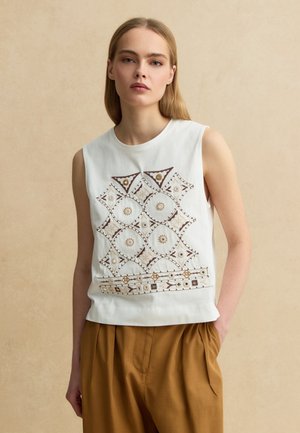 Femme portant un haut blanc sans manches avec une broderie géométrique marron et beige, associée à un pantalon ample marron, debout contre un mur beige.