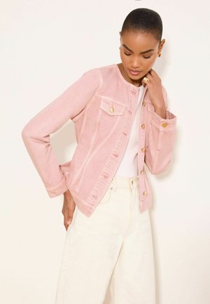 Kobieta w jasnoróżowej zapinanej na guziki kurtce na białej bluzce i kremowych spodniach z wysokim stanem, pozująca z jedną ręką na szyi, a drugą za sobą.