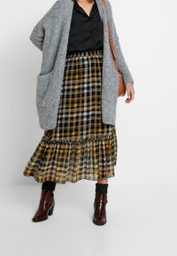 Gray knitted cardigan with pockets over a black blouse, paired with a yellow plaid pleated skirt and brown heeled boots.