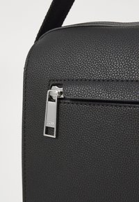 Sac en cuir noir texturé avec une tirette en métal argenté, de forme rectangulaire et un profil mince. Dispose d'une petite poche avant pour un rangement supplémentaire.