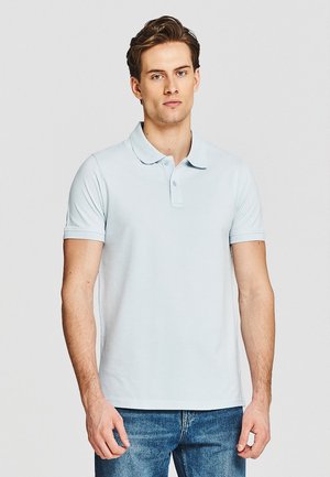 Młody mężczyzna w jasnoniebieskim polo z krótkim rękawem i niebieskich dżinsach stoi na tle jednolitego jasnego tła.