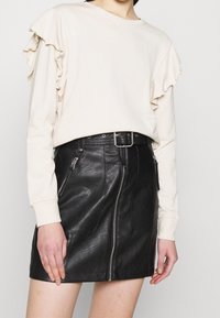 Top de manga larga color beige claro con detalles de volantes en las mangas, combinado con una mini falda de cuero negra que presenta cremalleras y un cinturón.