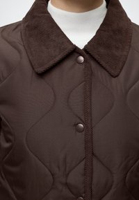 Veste marron matelassée avec col en velours côtelé et boutons-pression portée sur un top blanc à col montant, vue des épaules jusqu'au milieu de la poitrine.