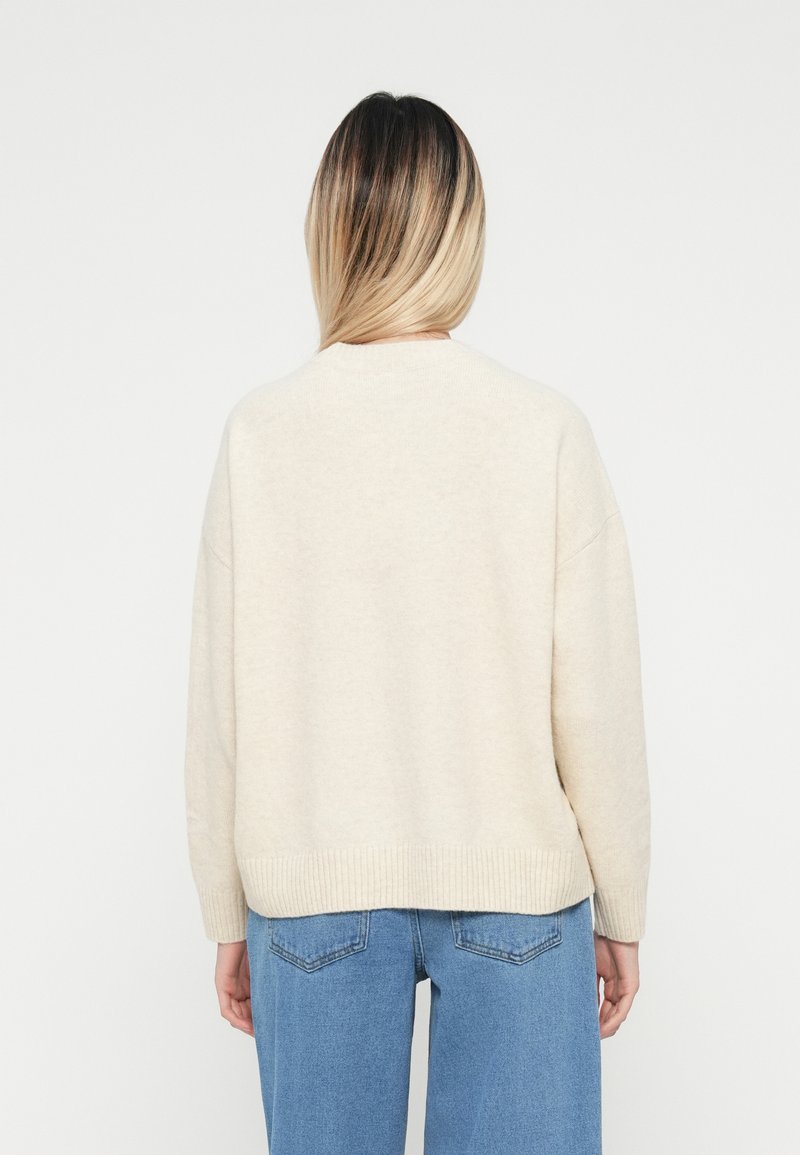 Osoba z ombre włosami nosi sweter w kolorze kremowym i niebieskie dżinsy, stojąc tyłem do kamery na tle gładkiej białej ściany.