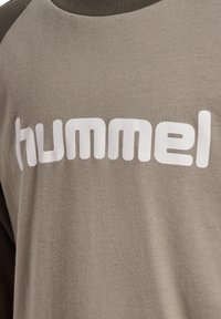 Grau und olivgrünes Baumwollhemd mit langen Ärmeln, das ein auffälliges weißes "hummel"-Logo auf der Brust präsentiert. Glatte Textur mit lockerer Passform.
