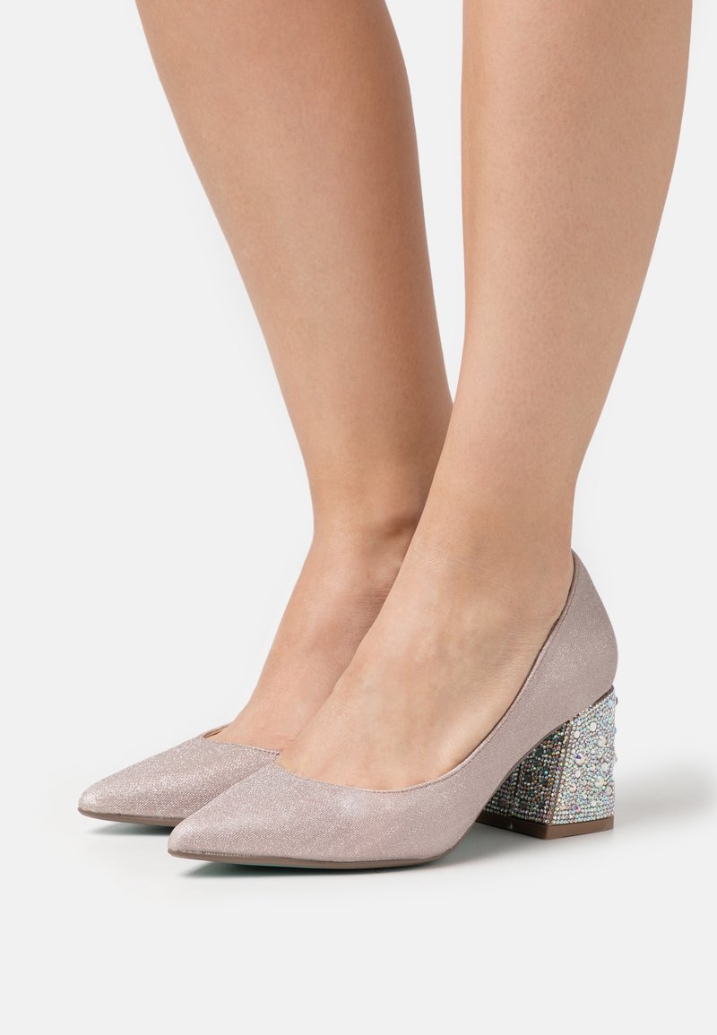 Femme portant des escarpins roses clairs à bouts pointus avec des talons blocs bas recouverts de petits strass brillants sur fond blanc.