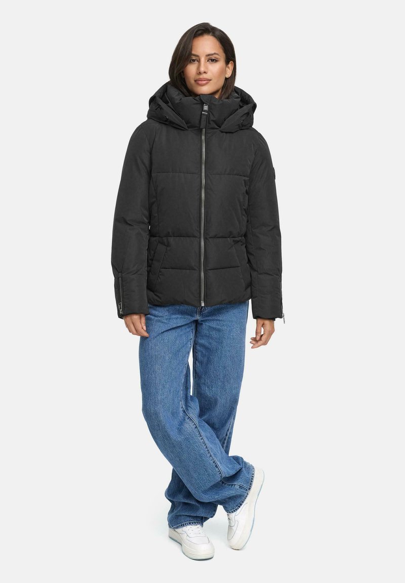 Zwarte pufferjack met een afneembare capuchon, ritssluiting aan de voorkant en een gewatteerd patroon, gecombineerd met lichtblauwe wijde jeans en witte sneakers.