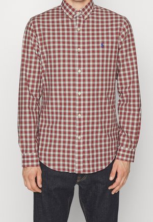 Mand iført en rød og hvid ternet skjorte med lange ærmer og mørke bukser, stående mod en ensfarvet baggrund.