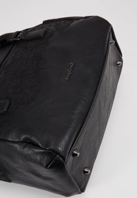 Sac en simili cuir noir avec un motif en dentelle délicat, fermeture éclair, accents métalliques et cinq pieds en métal au fond pour la stabilité.