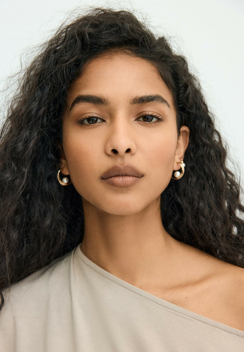 Jeune femme aux longs cheveux bouclés porte des boucles d’oreilles créoles dorées avec des perles et un haut clair à épaules dénudées sur un fond uni clair.