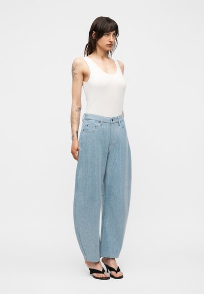 Γυναίκα με τατουάζ φοράει λευκό αμάνικο τοπ, ανοιχτό μπλε τζιν παντελόνι με φαρδιά μπατζάκια και μαύρα σανδάλια με τακούνι, στέκεται σε απλό φόντο.
