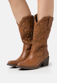 Stivali ankle in pelle marrone con intricati motivi di cucitura, punta affilata e tacco basso. Presentano una chiusura con zip sul lato.