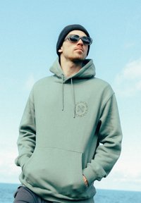 Haut à capuche vert menthe avec une poche avant, logo sur la poitrine, cordons au cou, associé à un bonnet en tricot sombre et des lunettes de soleil.