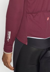 Weinrotes Radtrikot mit langen Ärmeln, das ein Polartec-Label am linken Arm trägt. Es ist mit schwarzen Fahrradshorts kombiniert.