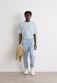 Lys blå ribbet kortermet skjorte parret med lys blå joggebukser. Modellen holder en beige hettegenser. Hvite sneakers fullfører antrekket.