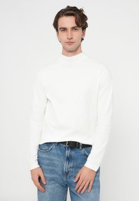 Biała koszula z długim rękawem i wysokim kołnierzem, noszona z niebieskimi dżinsami denimowymi, z czarnym paskiem i luźnym krojem. Gładka tekstura.