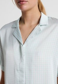 Blusa verde chiaro a maniche corte con colletto, presenta un motivo ripetuto di piccoli punti rosa, texture morbida e vestibilità comoda.