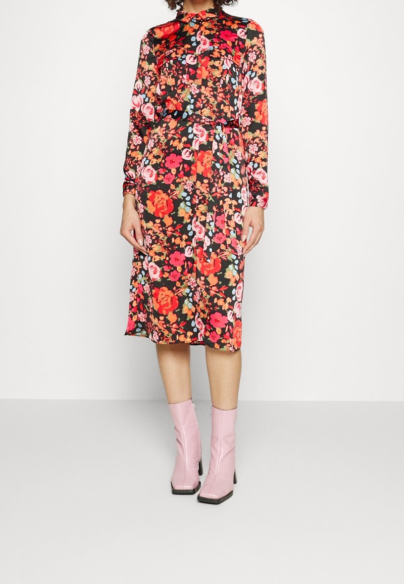 Femme portant une robe noire à manches longues et longueur genou avec un imprimé florale rouge et rose vif, associée à des bottines à talons rose clair.