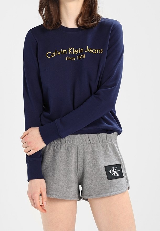 Námořnická modrá tričko s dlouhým rukávem s nápisem "Calvin Klein Jeans od roku 1978" ve žluté barvě. Šedé šortky s černou nášivkou se logem. Pohodlný bavlněný materiál.
