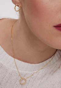 Złote kolczyki-hoop z małymi perłami, połączone z delikatnym złotym naszyjnikiem łańcuszkowym z okrągłym wisiorkiem. Tło z białym swetrem dzianinowym.