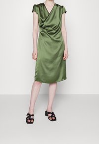 Robe satin olive à mi-mollet avec un décolleté drapé et des manches courtes. Associée à des sandales noires à talons carrés et à lanières.
