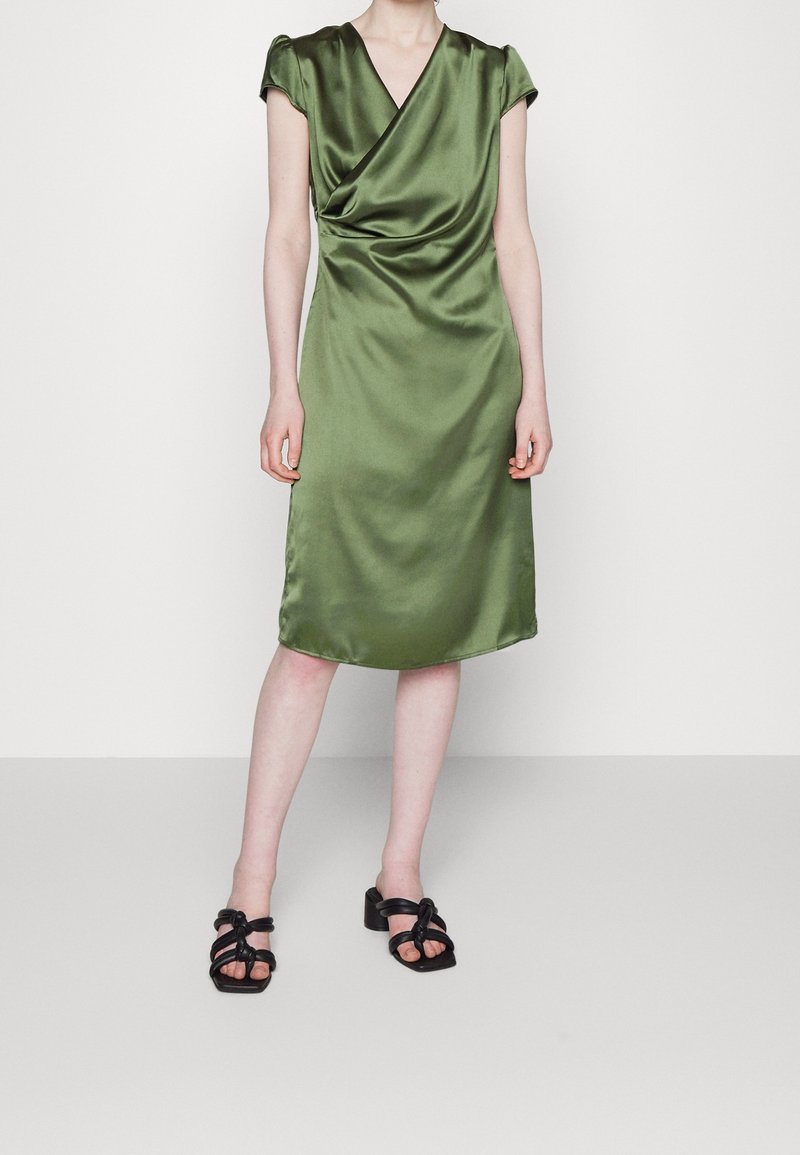 Robe satin olive à mi-mollet avec un décolleté drapé et des manches courtes. Associée à des sandales noires à talons carrés et à lanières.