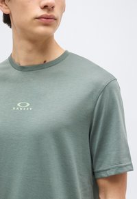 Νέος άνθρωπος που φοράει μια μονόχρωμη πράσινη μπλούζα Oakley με κοντά μανίκια και στρογγυλή λαιμόκοψη, με φόντο ένα απλό φωτεινό φόντο.