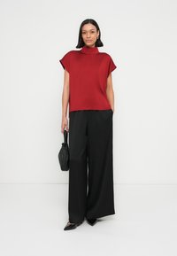 Czerwony top z krótkim rękawem i wysokim kołnierzem, zestawiony z czarnymi spodniami o szerokich nogawkach; modelka trzyma małą czarną torebkę i nosi czarne buty na szpic.