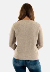 Maglione beige a costine con maniche lunghe e scollatura rotonda, realizzato in materiale morbido. Indossato con jeans blu con tasche posteriori distintive.