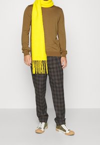 Bruine trui, gele franje-sjaal, geruite broek in donkere tinten en groene sneakers met witte accenten, gepresenteerd in een staande pose.