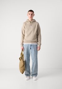 Bež kapuca s vrvico in logotipom, svetlo denim jeans, drži svetlo rjavo mehko torbo z naramnicami in nosi bele čevlje.