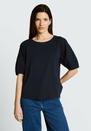 Femme aux cheveux raides et foncés portant un haut noir à manches courtes avec des manches en dentelle et un jean bleu, debout avec une main sur la hanche.