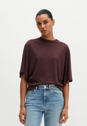 Frau mit dunklem, zurückgebundenem Haar, die ein lockeres dunkelbraunes Kurzarmshirt und hellblaue Jeans mit hoher Taille vor einem einfarbigen Hintergrund trägt.