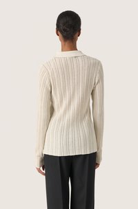 Donna con i capelli castani raccolti in un chignon, vestita con un top a maniche lunghe di maglia a coste color crema e pantaloni neri, vista di spalle su uno sfondo neutro.