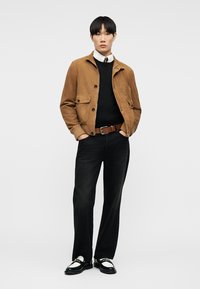 Young man wearing a tan suede jacket, black sweater, white shirt with tie, black pants, brown belt, and black-and-white loafers, standing with hands in pockets.