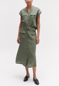 Vestido de lino verde con cuello, mangas cortas y botones frontales. Presenta dos bolsillos en el pecho y un cinturón de tela con una hebilla redonda.