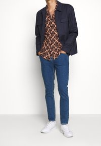 Veste bleu marine portée sur une chemise à motifs marron-orange, associée à un jean bleu et des baskets blanches. Caractéristiques : deux poches avant sur la veste.