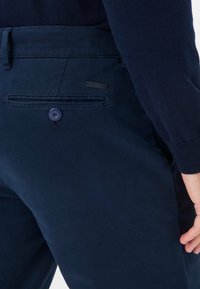 Pantaloni blu navy realizzati in tessuto misto cotone con una texture liscia. Presentano una tasca posteriore con bottone e un'etichetta metallica decorativa.