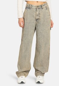 Persona vistiendo jeans holgados de lavado claro con patas anchas, zapatillas blancas y un top corto de manga larga frente a un fondo liso.