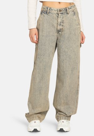 Persoon in losse, lichtgewassen baggy jeans met wijde pijpen, witte sneakers en een korte lange mouw top tegen een eentonige achtergrond.