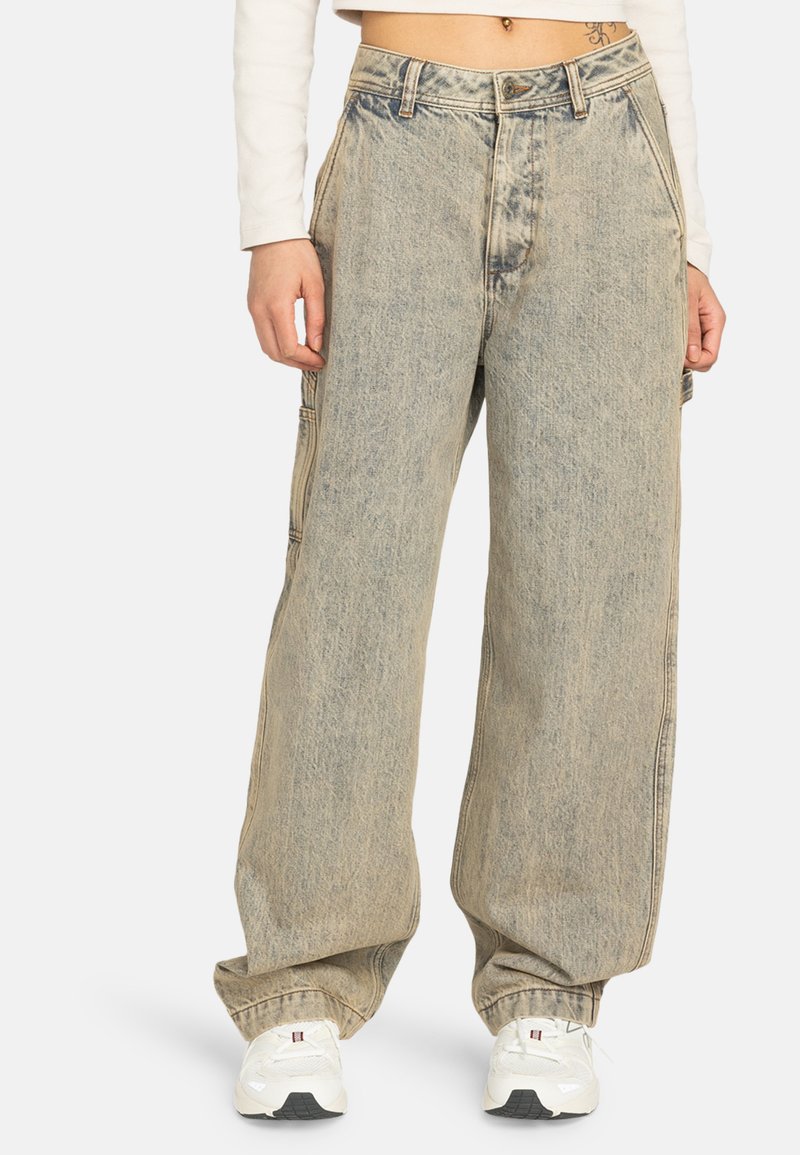Persona vistiendo jeans holgados de lavado claro con patas anchas, zapatillas blancas y un top corto de manga larga frente a un fondo liso.