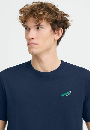 Junger Mann mit lockigem braunem Haar, der ein marineblaues T-Shirt mit einem kleinen grünen Krokodil-Logo auf der linken Brust trägt.