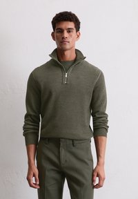 Grüner Strickpullover mit einem Reißverschluss am Hals und gerippten Manschetten, kombiniert mit passenden grünen Hosen. Die Textur hat ein leichtes Muster.