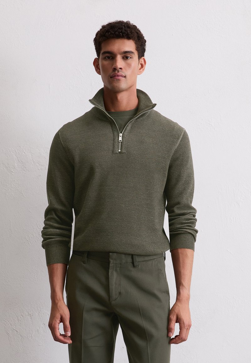 Grüner Strickpullover mit einem Reißverschluss am Hals und gerippten Manschetten, kombiniert mit passenden grünen Hosen. Die Textur hat ein leichtes Muster.