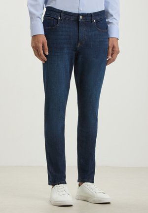 Jeans in denim blu scuro con vestibilità slim, dotati di tasche frontali e dettagli di whiskering leggeri, abbinati a sneakers bianche.